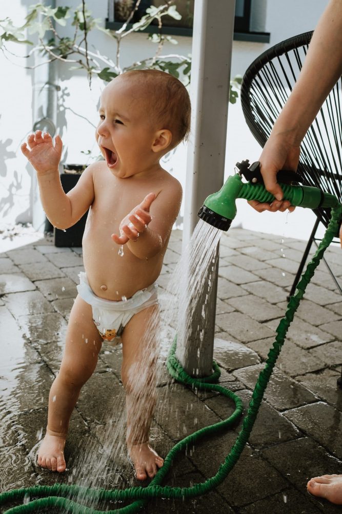 Baby in Windeln wird von Gartenschlauch naß gespritzt