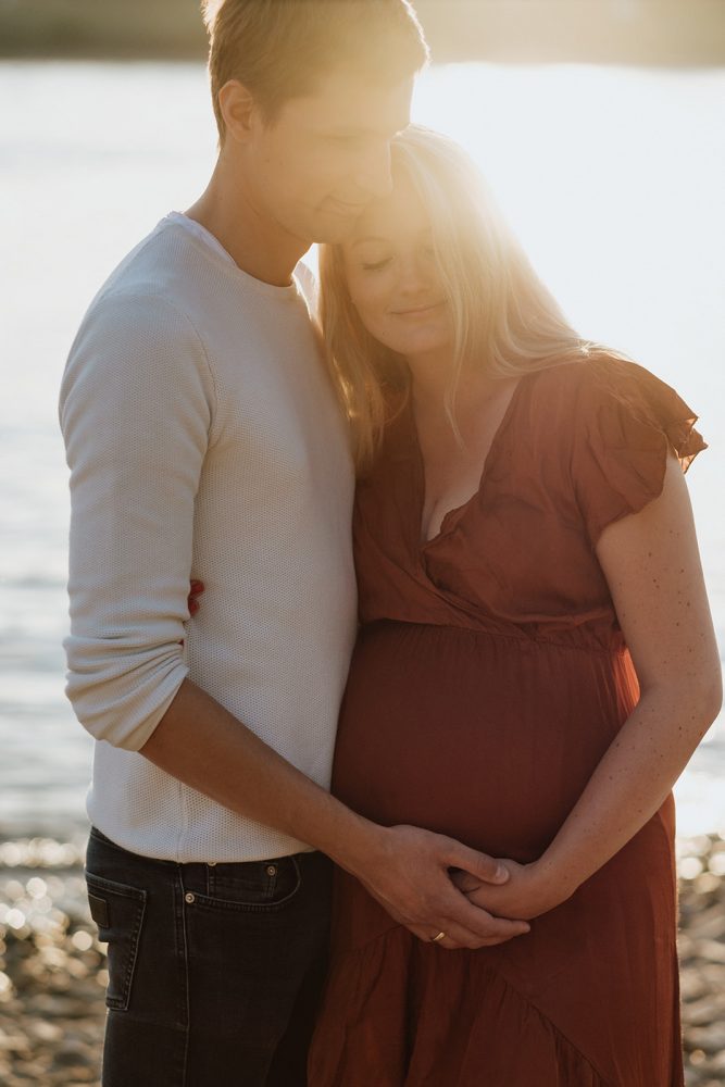 Portrait von einem Pärchen im Gegenlicht vor einem Fluss während eines Babybauchshootings